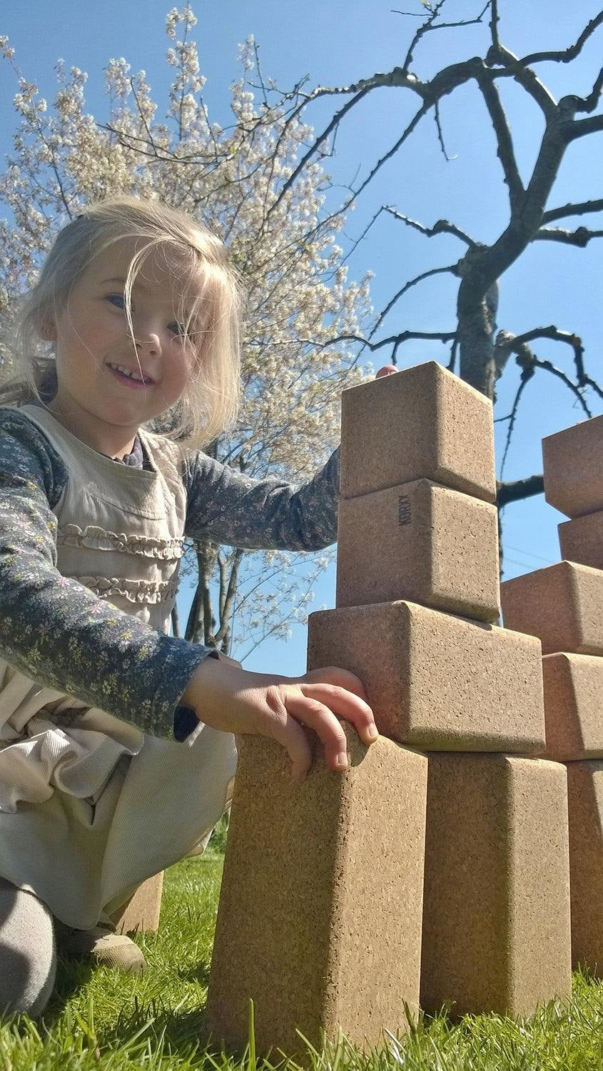 A young girl stacking large, natural cork Korxx building blocks outdoors on a sunny day.