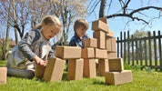 Two children building a large tower with oversized, natural cork blocks on a sunny green lawn.