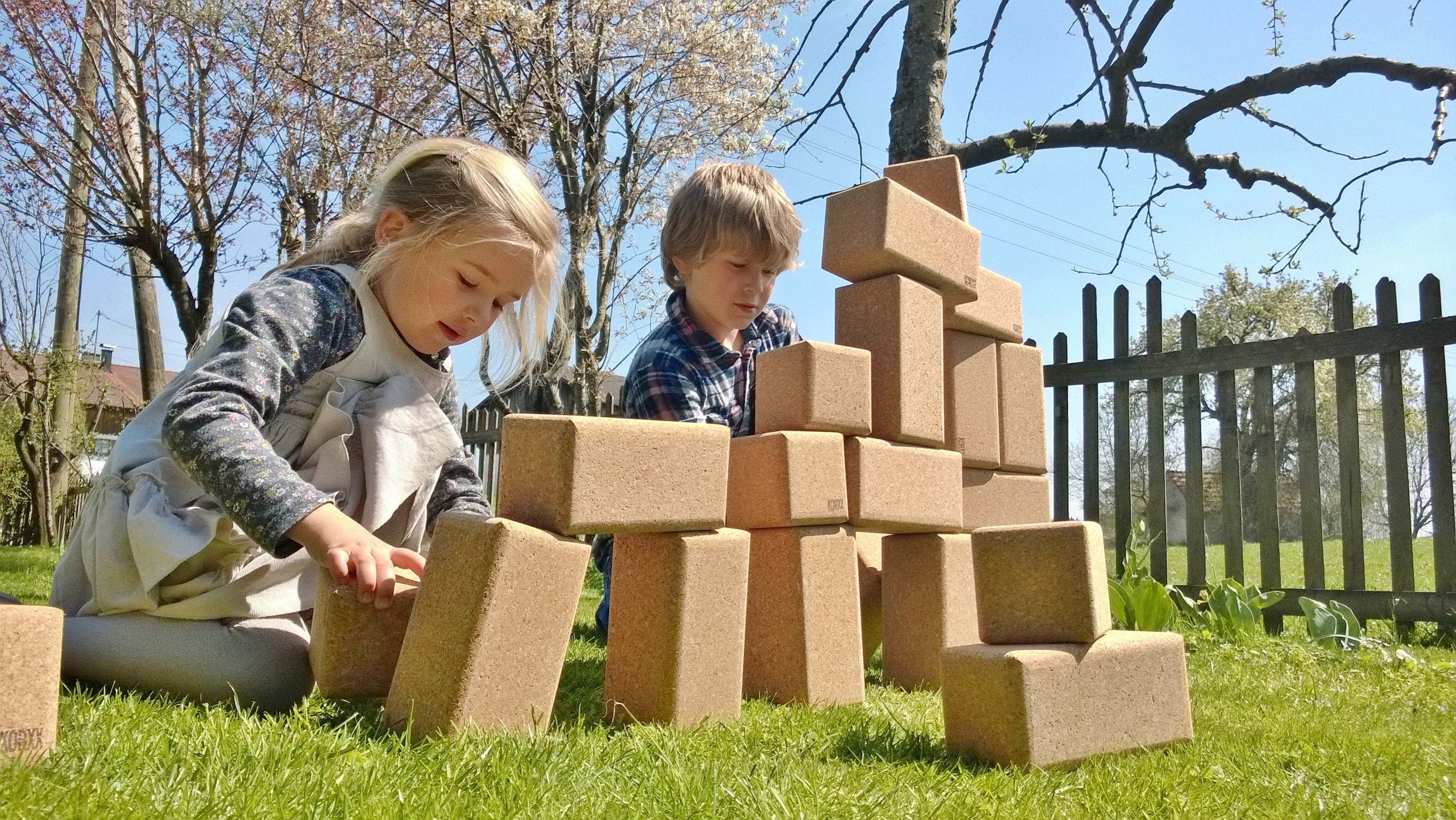 Two children building a large tower with oversized, natural cork blocks on a sunny green lawn.