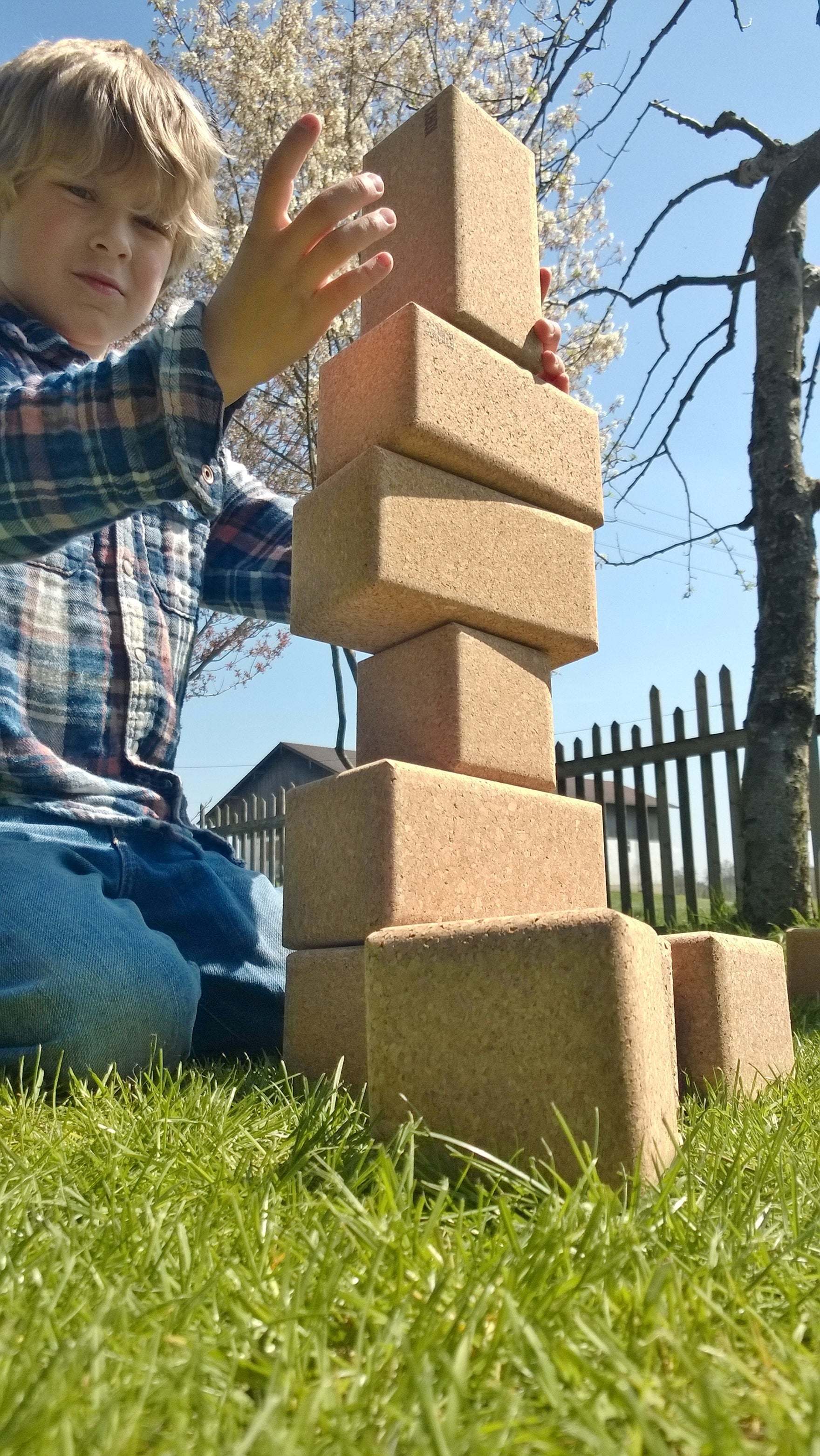 A young boy stacking large, natural cork building blocks into a tall tower on a sunny outdoor lawn.