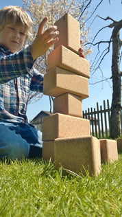 A young boy stacking large, natural cork building blocks into a tall tower on a sunny outdoor lawn.