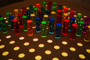 Vibrant acrylic rainbow pegs inserted into a natural birch grid pegboard illuminated from below.