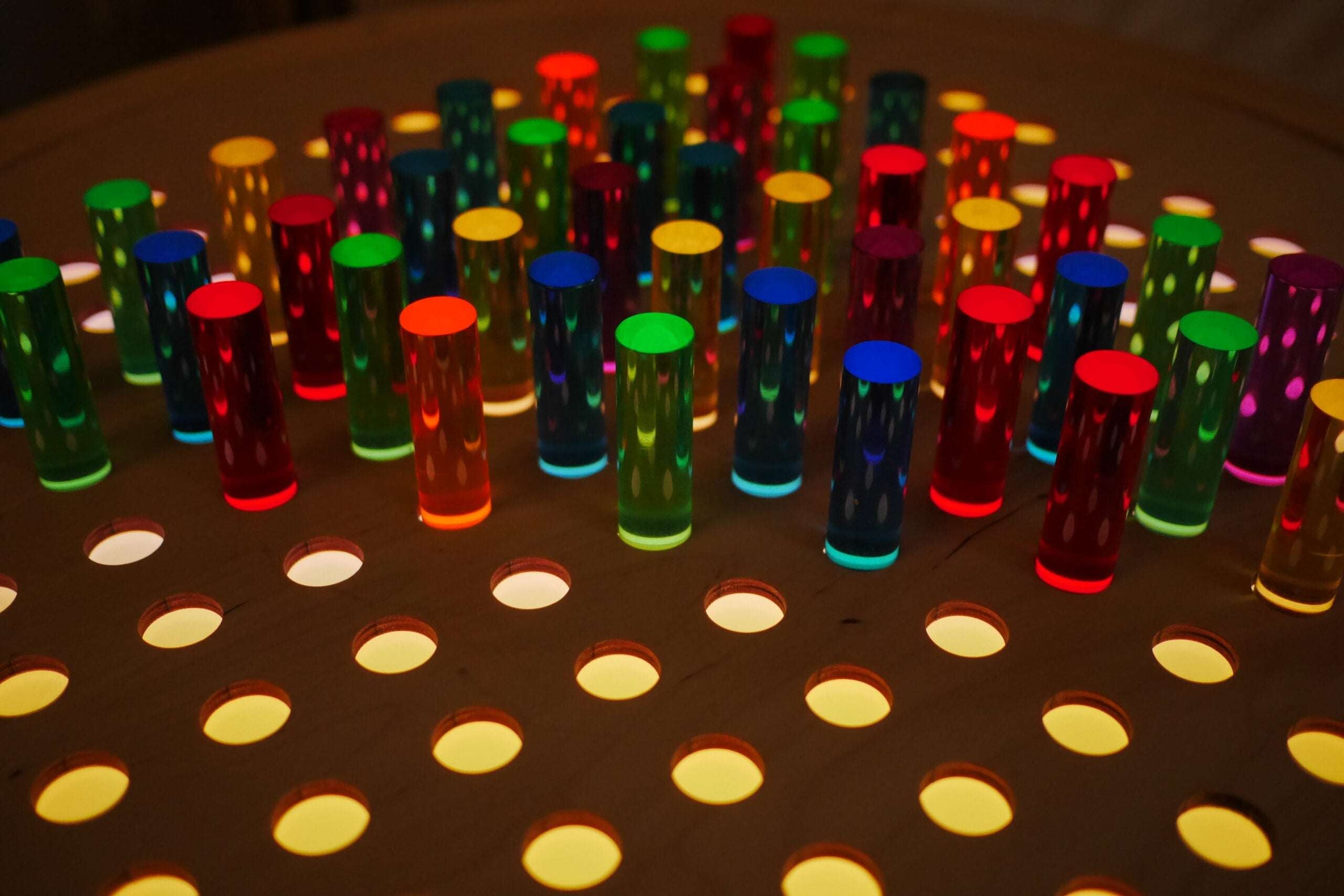 Vibrant acrylic rainbow pegs inserted into a natural birch grid pegboard illuminated from below.