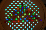 Circular birch pegboard with glowing, colorful acrylic pegs arranged in a grid pattern on a light table.