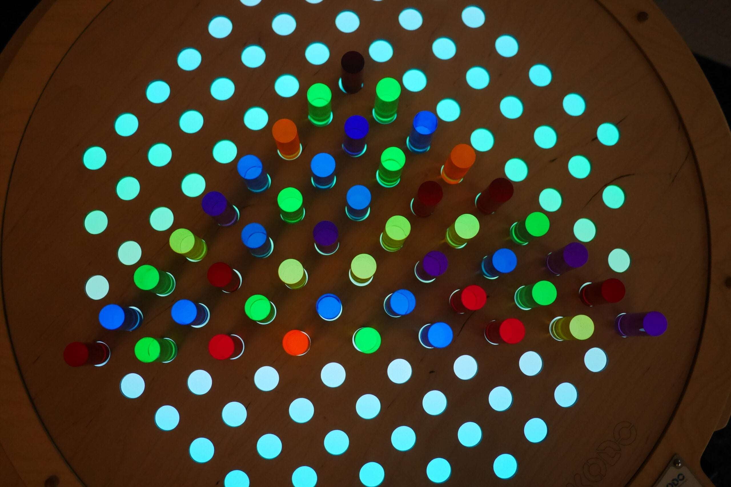 Circular birch pegboard with glowing, colorful acrylic pegs arranged in a grid pattern on a light table.