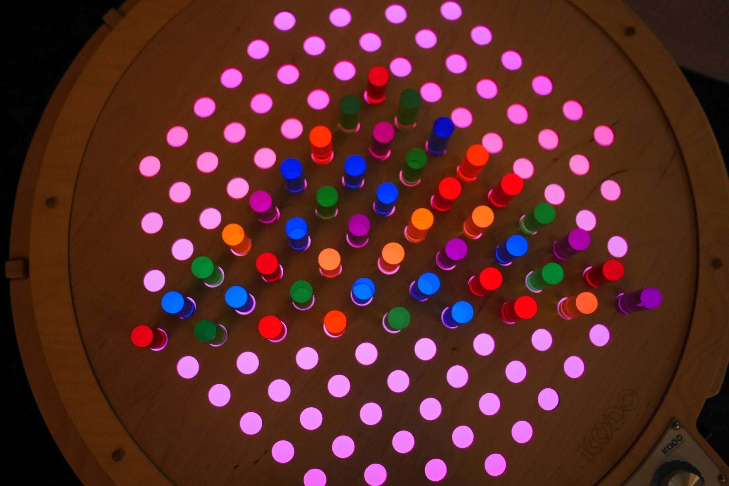 Circular birch pegboard with glowing, colorful acrylic pegs arranged in a grid pattern on a light table.