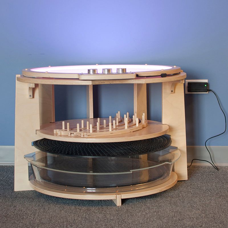 Light wood storage pod with a circular light table top, featuring organized shelves for trays, boards, and accessories.
