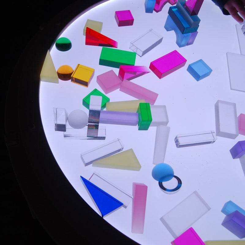 Colorful, frosted translucent building blocks in various geometric shapes arranged on a circular glowing light table.