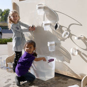 Children playing with a magnetic water circuit wall featuring clear tubes, white cups, and a hand pump on a wooden panel.