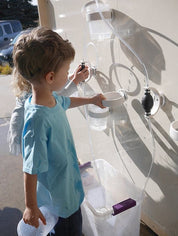 Children playing with a magnetic wall water circuit featuring clear tubes, white funnels, and pumps on a vertical surface.