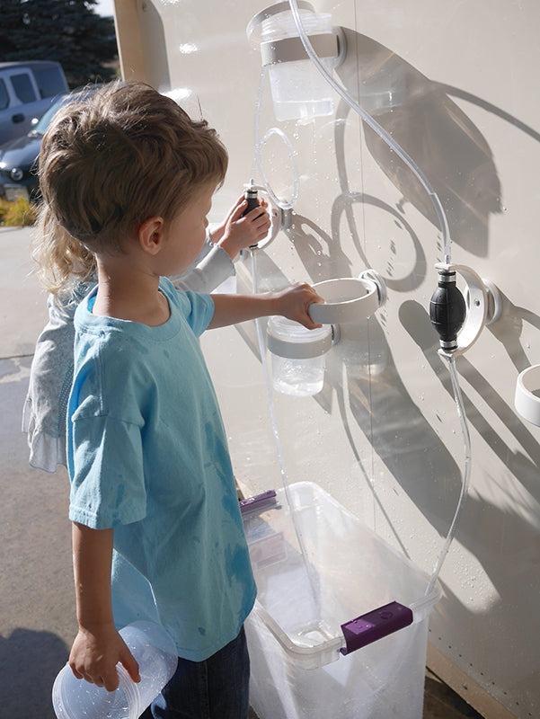 Children playing with a magnetic wall water circuit featuring clear tubes, white funnels, and pumps on a vertical surface.