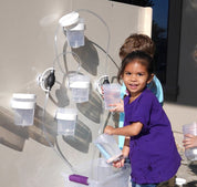 Children playing with a magnetic wall water circuit featuring clear plastic cups, tubing, and pumps on a beige panel.
