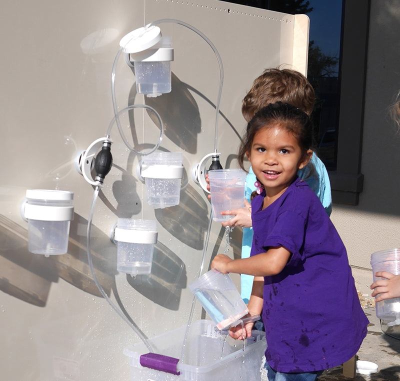 Children playing with a magnetic wall water circuit featuring clear plastic cups, tubing, and pumps on a beige panel.