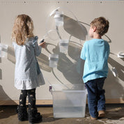 Children playing with a magnetic wall water circuit featuring clear tubes, white modular cups, and a base water collection bin.