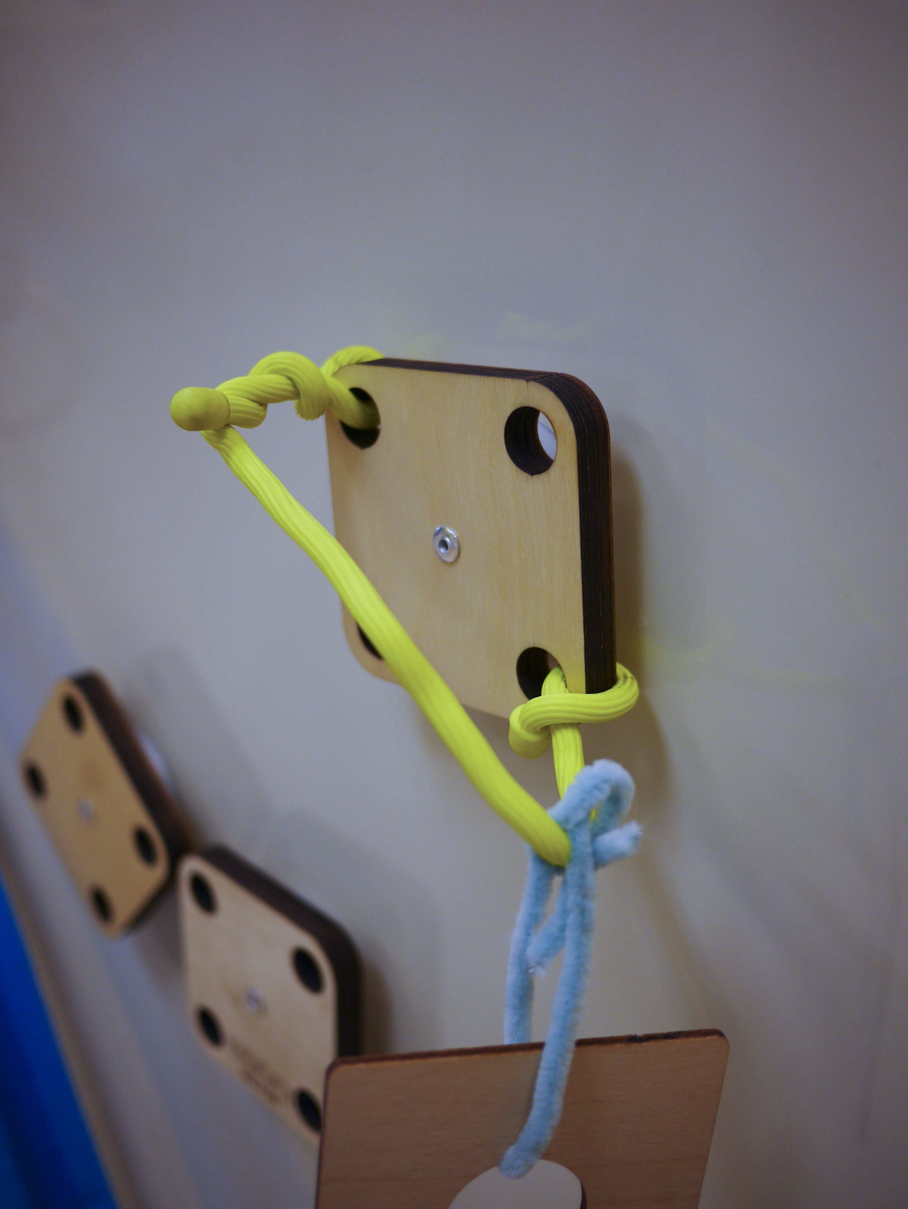 Square wooden mounting platform with yellow cord and blue pipe cleaner attached to a magnetic wall.
