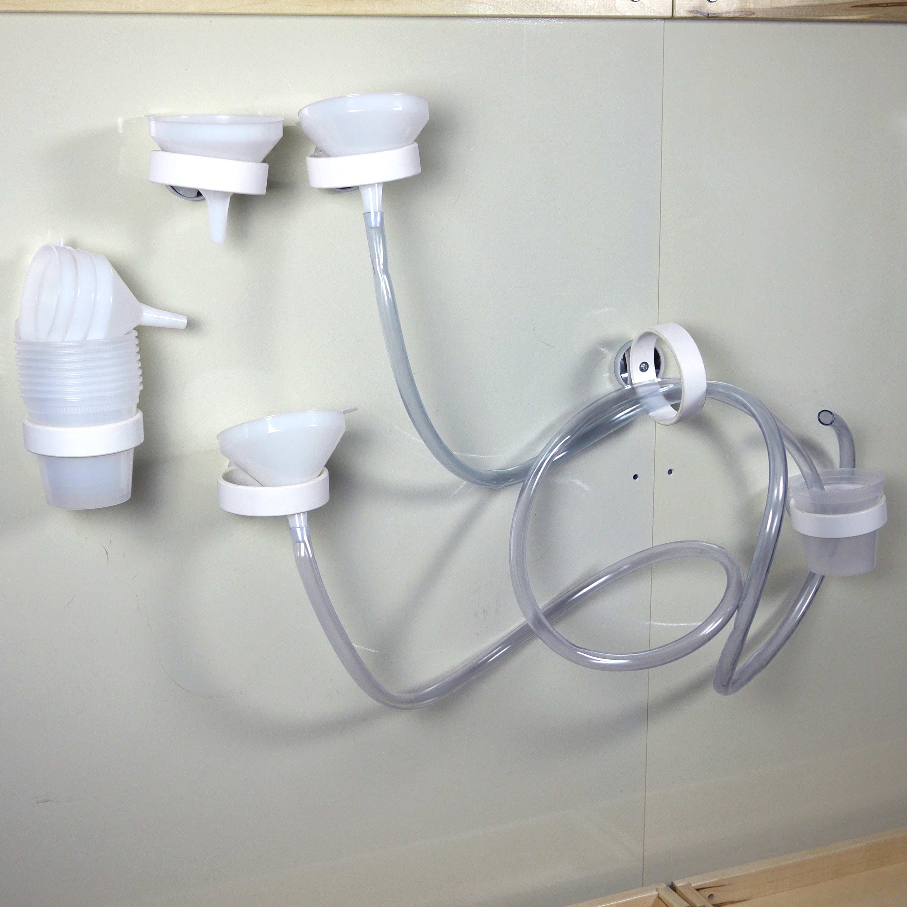 White magnetic funnels, clear tubing, and cups attached to a wall for interactive outdoor water play.