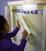 Child placing a white plastic ramp on a magnetic wall featuring blue word and symbol magnets.