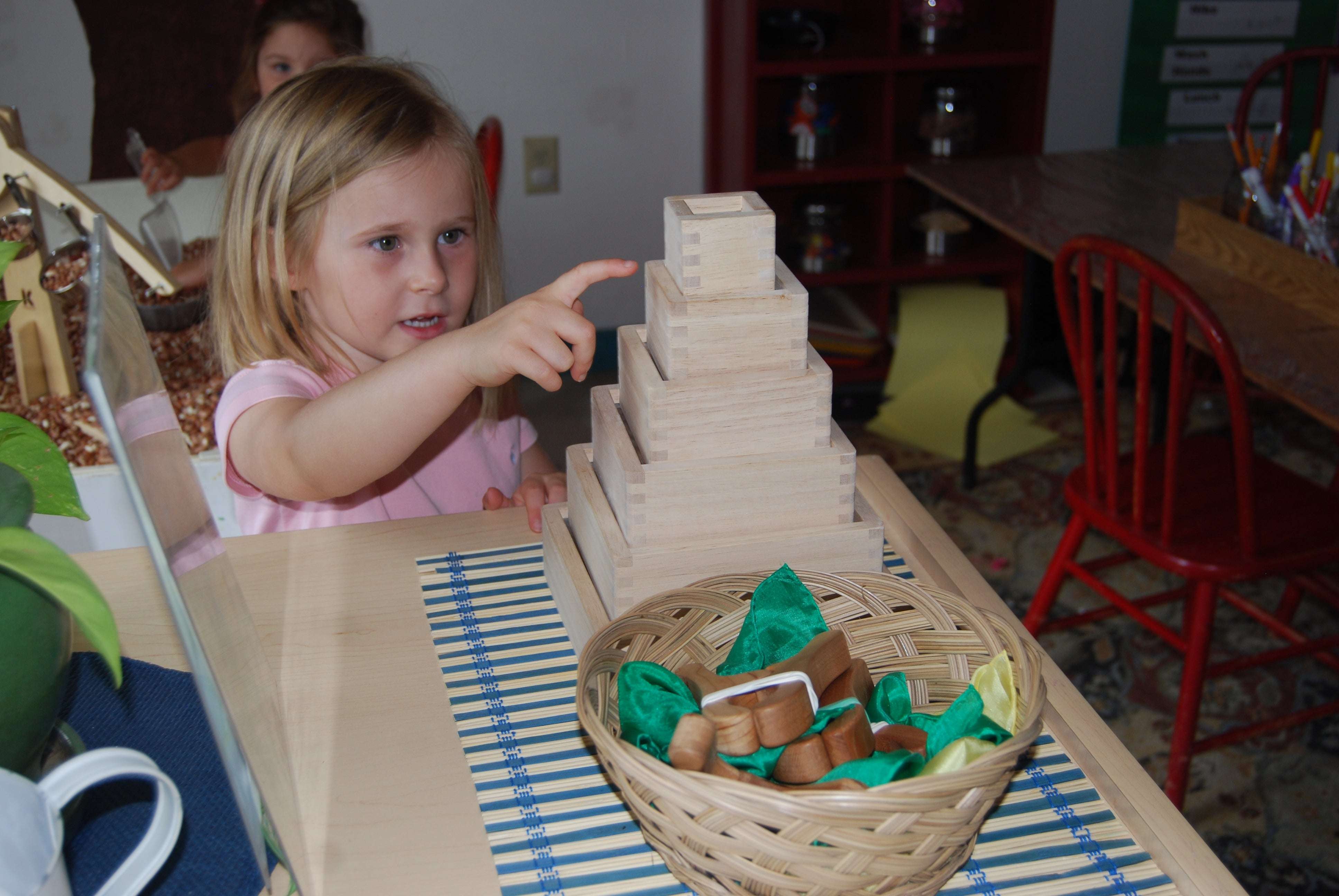 A young girl points at a stack of six natural hardwood magnifying blocks with inset acrylic windows on a classroom table.