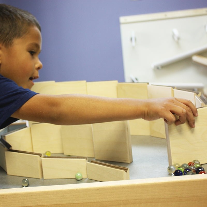 Child building a wooden marble maze ramp with colorful glass marbles on a table.