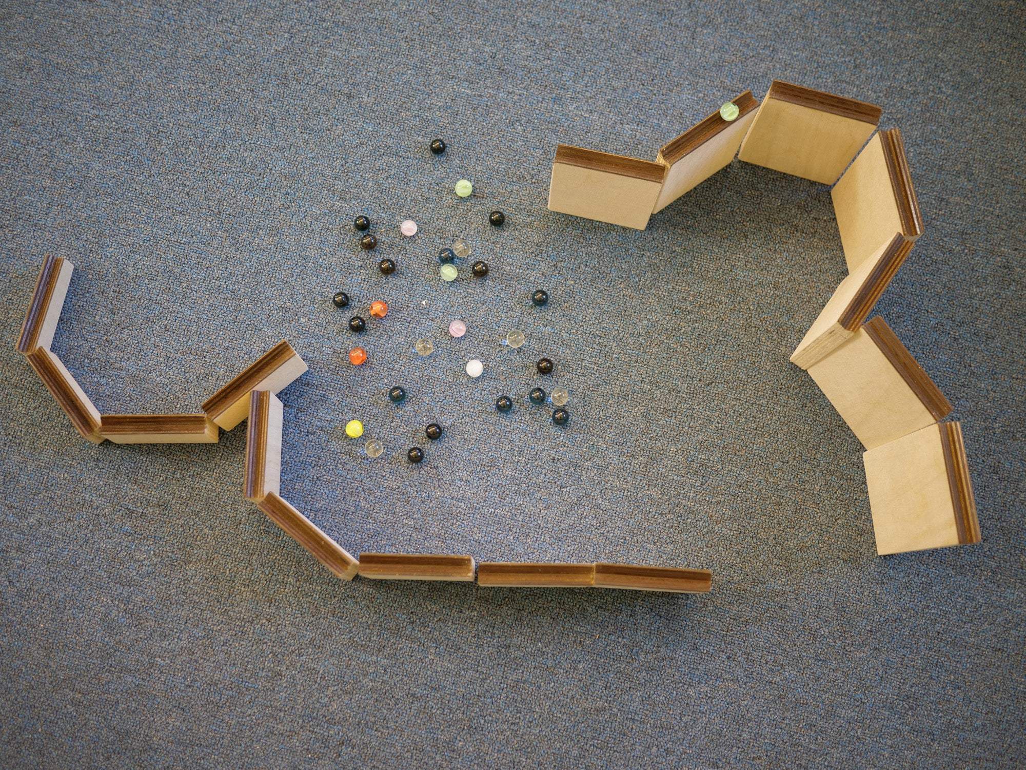 Wooden marble maze ramps arranged on a blue carpet with a collection of colorful glass marbles in the center.