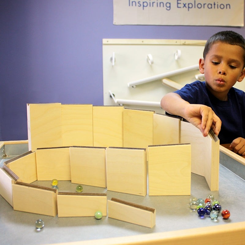 Child building a custom wooden marble maze ramp with colorful glass marbles on a magnetic play surface.