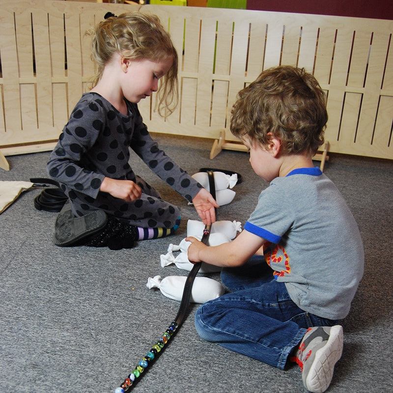 Two children building a marble track with a flexible black rubber ramp and small white weighted bags on a grey carpet.