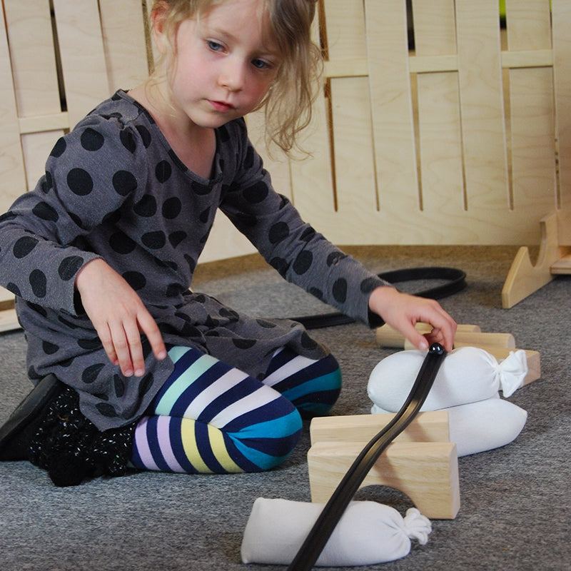 A child building a marble track using a flexible black rubber ramp supported by wooden blocks and white sandbags.