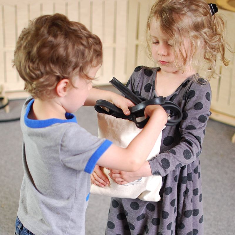 Two children holding a beige canvas bag filled with flexible black rubber tracks for a marble run STEM toy.