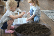 Two children playing with soil in a large, round, clear plastic Messy Material Tray on a wooden floor.