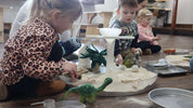 Children playing with a clear, round Messy Material Tray filled with sand, felt dinosaurs, and sensory toys on a floor.