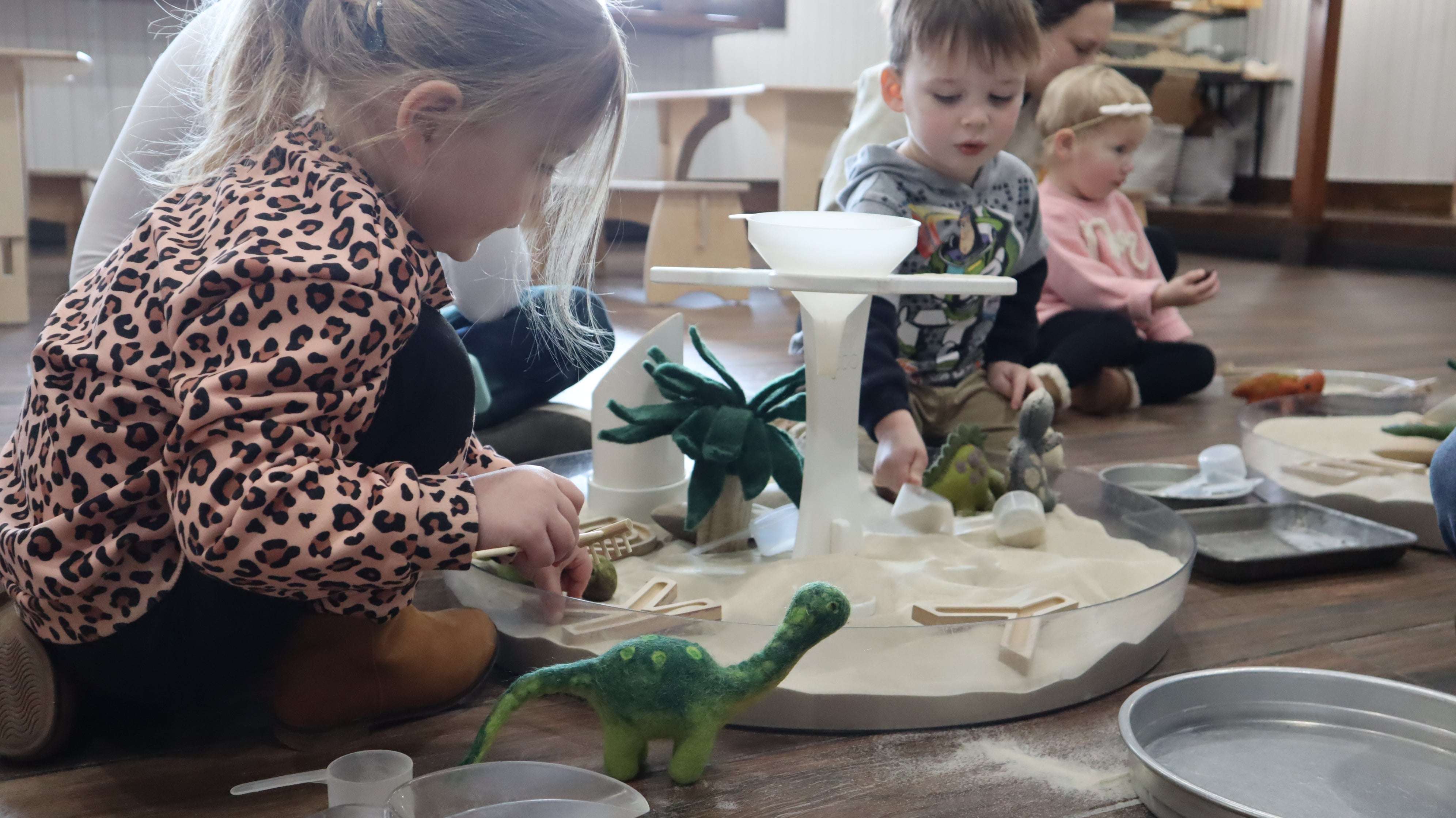 Children playing with a clear, round Messy Material Tray filled with sand, felt dinosaurs, and sensory toys on a floor.