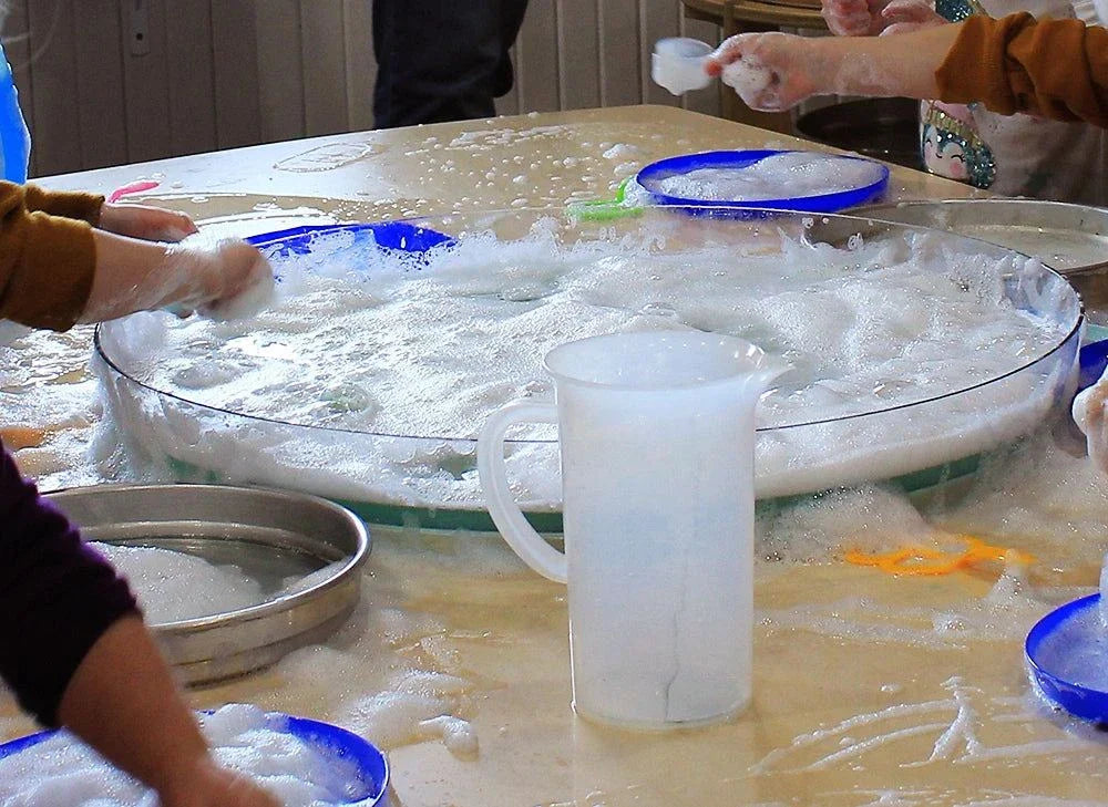 Clear, circular Messy Material Tray filled with soapy water and foam for sensory play on a classroom table.