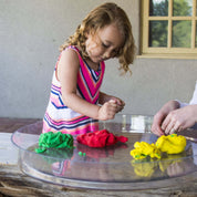 Child playing with green, red, and yellow modeling clay on a clear, round, shallow Messy Material Tray.