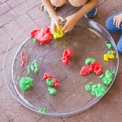 Child playing with colorful modeling clay on a large, round, clear plastic messy material tray on a brick patio.