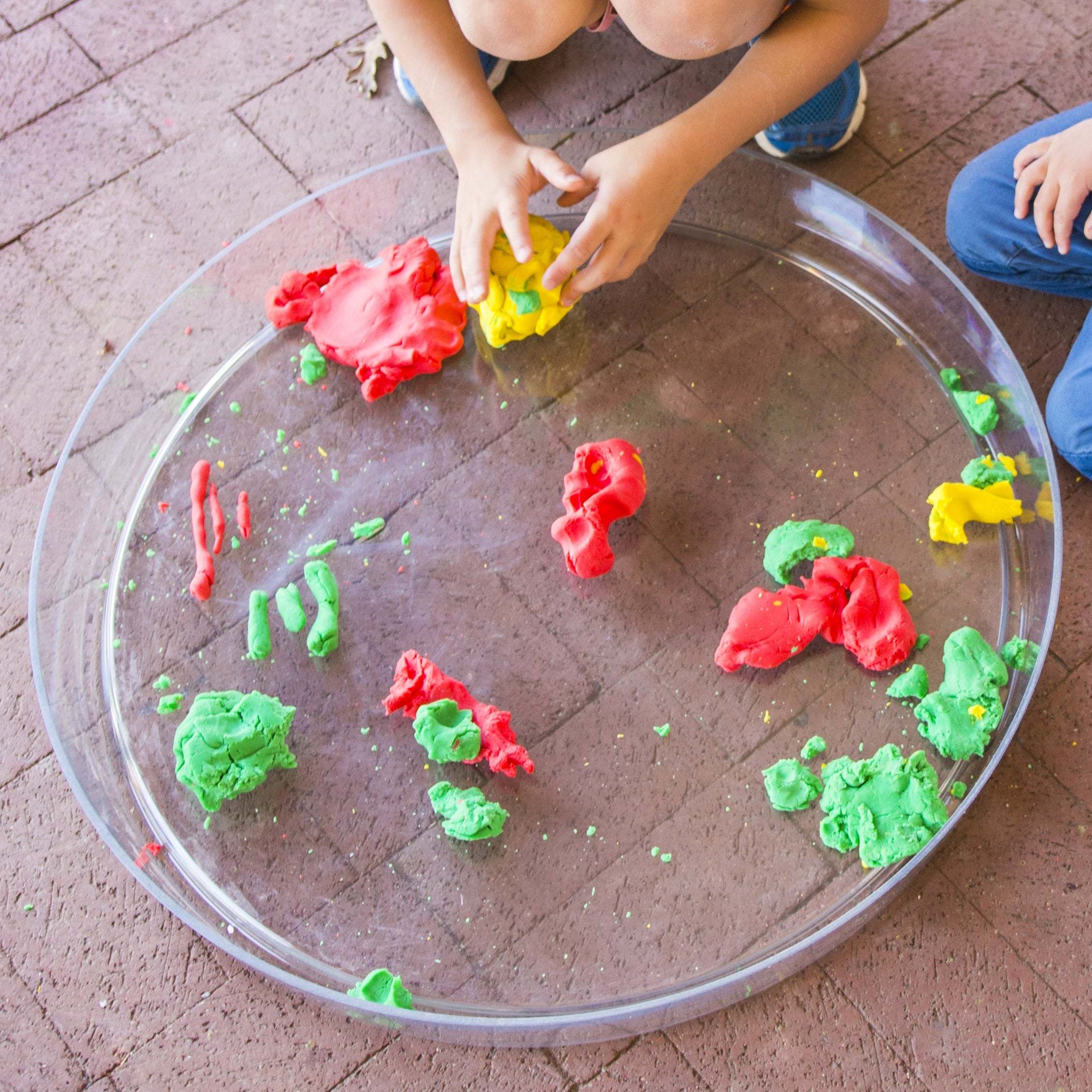 Child playing with colorful modeling clay on a large, round, clear plastic messy material tray on a brick patio.