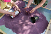 Children playing with shiny stainless steel metal cups and purple sand in a wooden sensory tray.