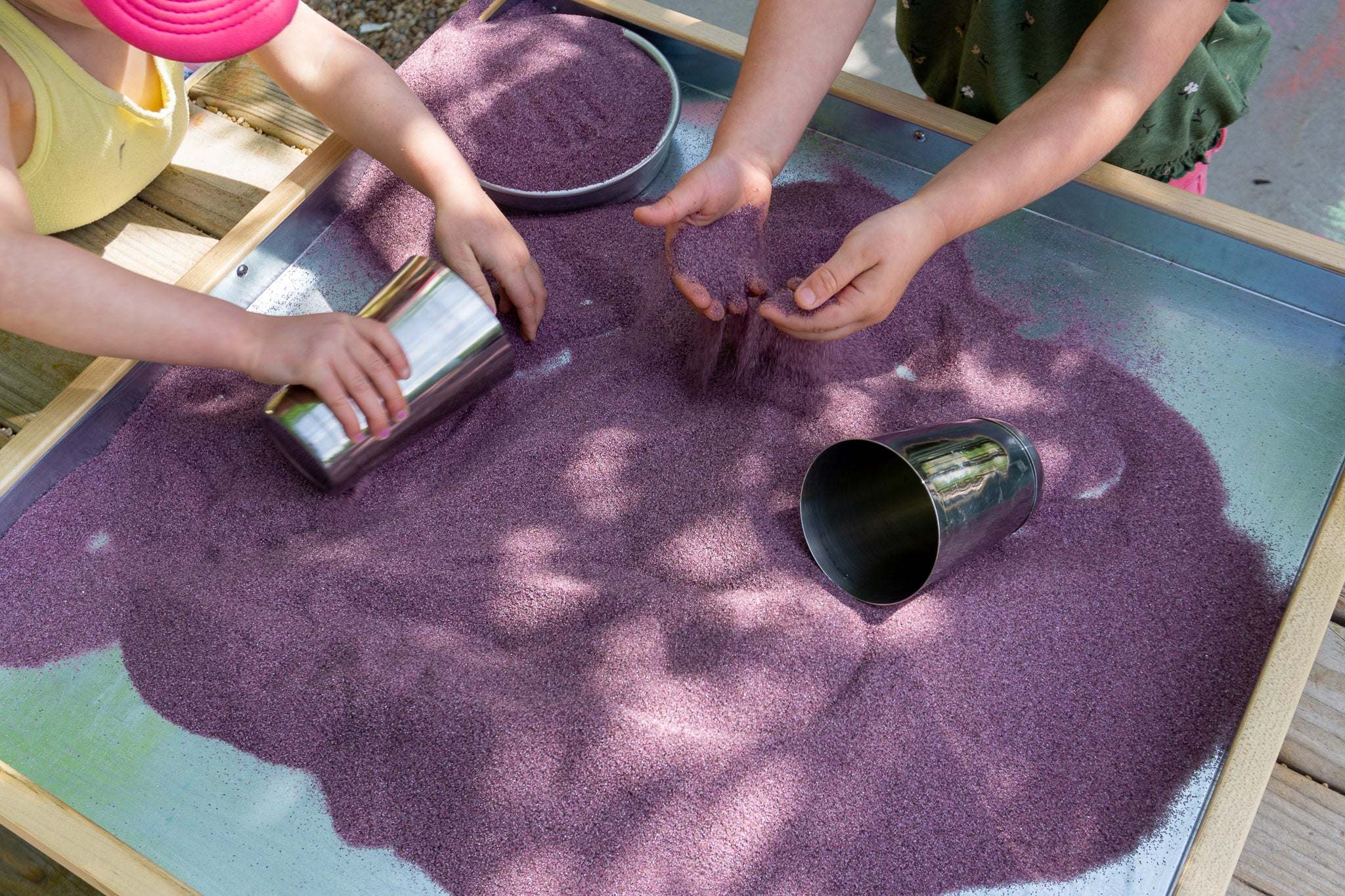 Children playing with shiny stainless steel metal cups and purple sand in a wooden sensory tray.