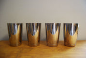 Four shiny, polished stainless steel cups lined up on a wooden surface against a neutral wall.