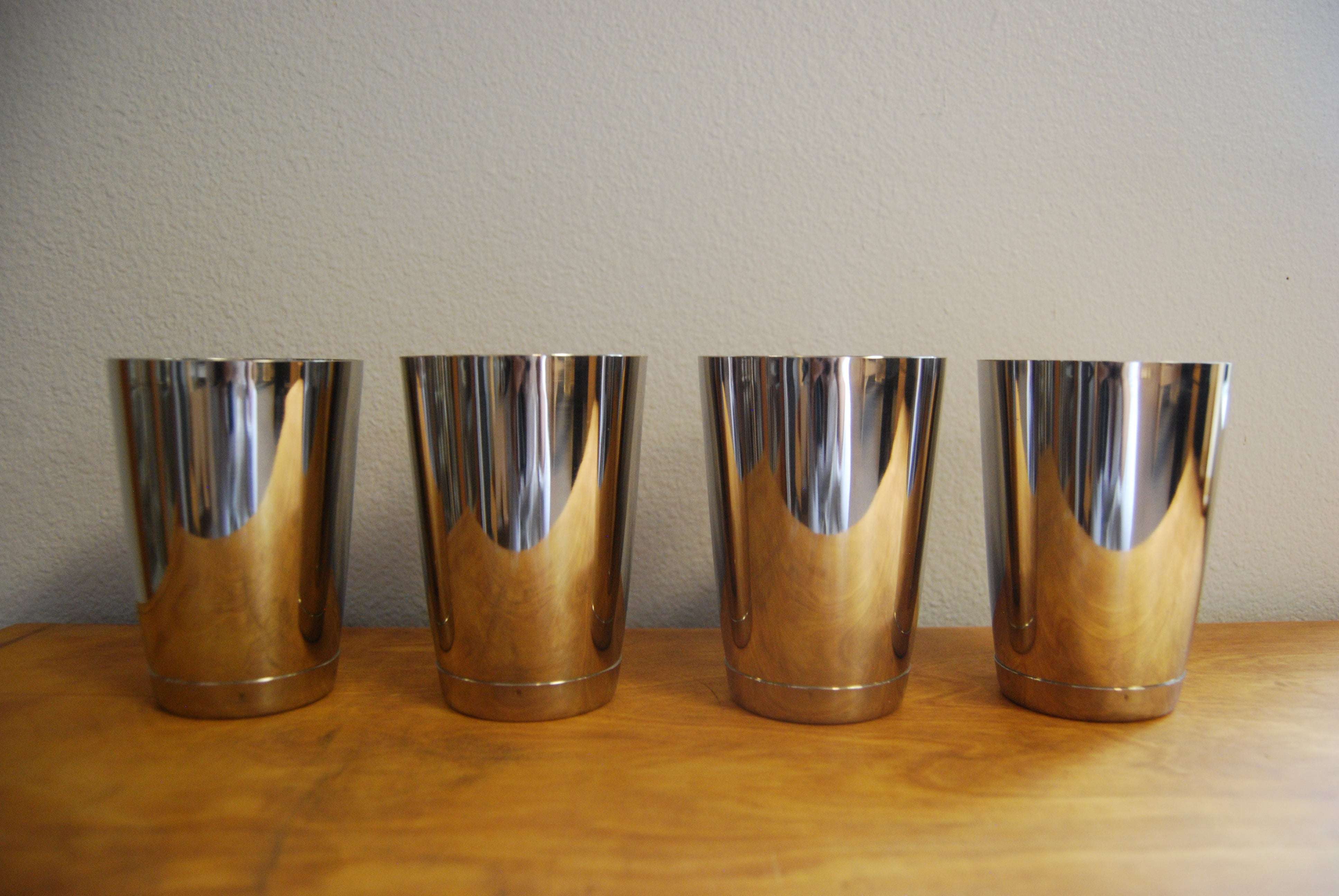 Four shiny, polished stainless steel cups lined up on a wooden surface against a neutral wall.