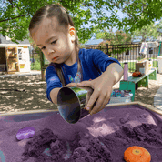 Young girl using a shiny, cylindrical metal cup to mold purple kinetic sand on an outdoor play table.