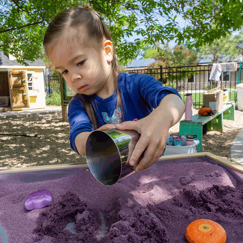Young girl using a shiny, cylindrical metal cup to mold purple kinetic sand on an outdoor play table.