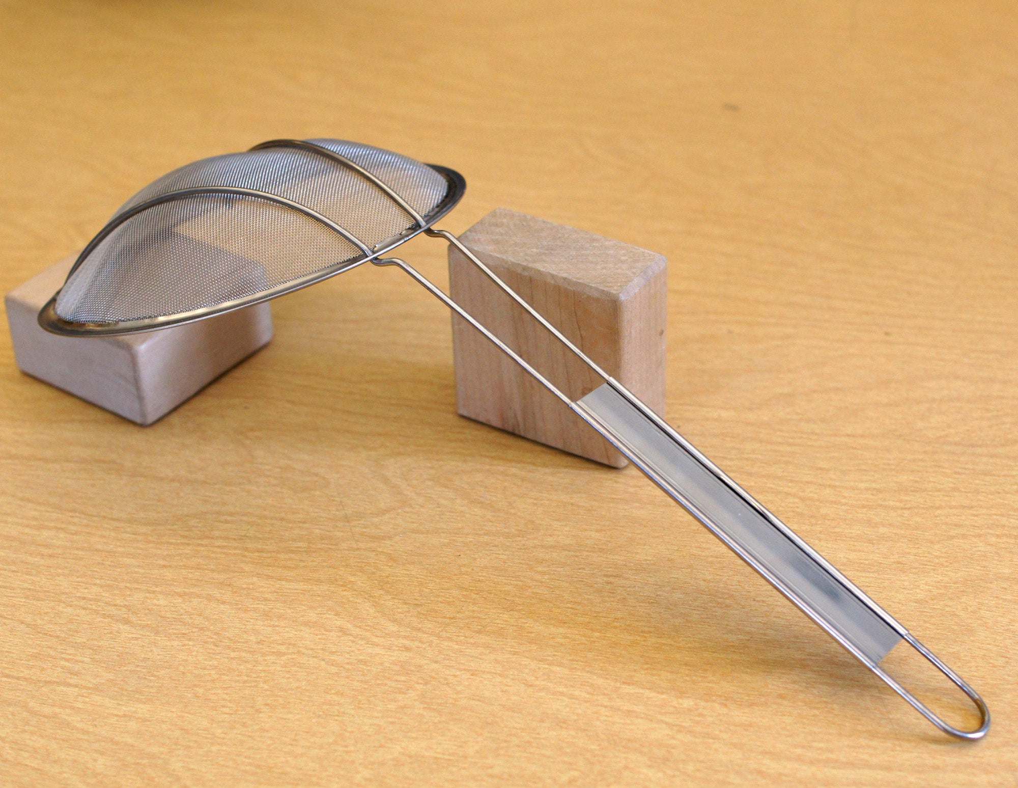Fine-mesh metal sand sifter with a long stainless steel handle resting on two small wooden blocks.