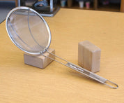 Round stainless steel mesh sand sifter with a long handle, resting on a wooden block on a light wood table.