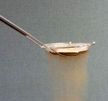 Fine-mesh metal sand sifter with a long handle, straining golden sand against a grey textured background.