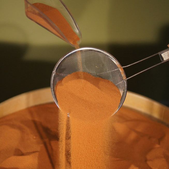 Fine-mesh metal sand sifter straining orange Jurassic sand into a wooden bowl.