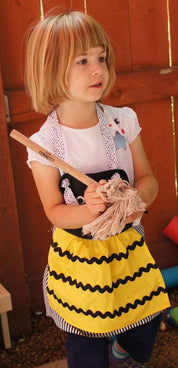 Young girl holding a mini mop with a wooden handle and soft cotton head, wearing a yellow and black bumblebee-style apron.