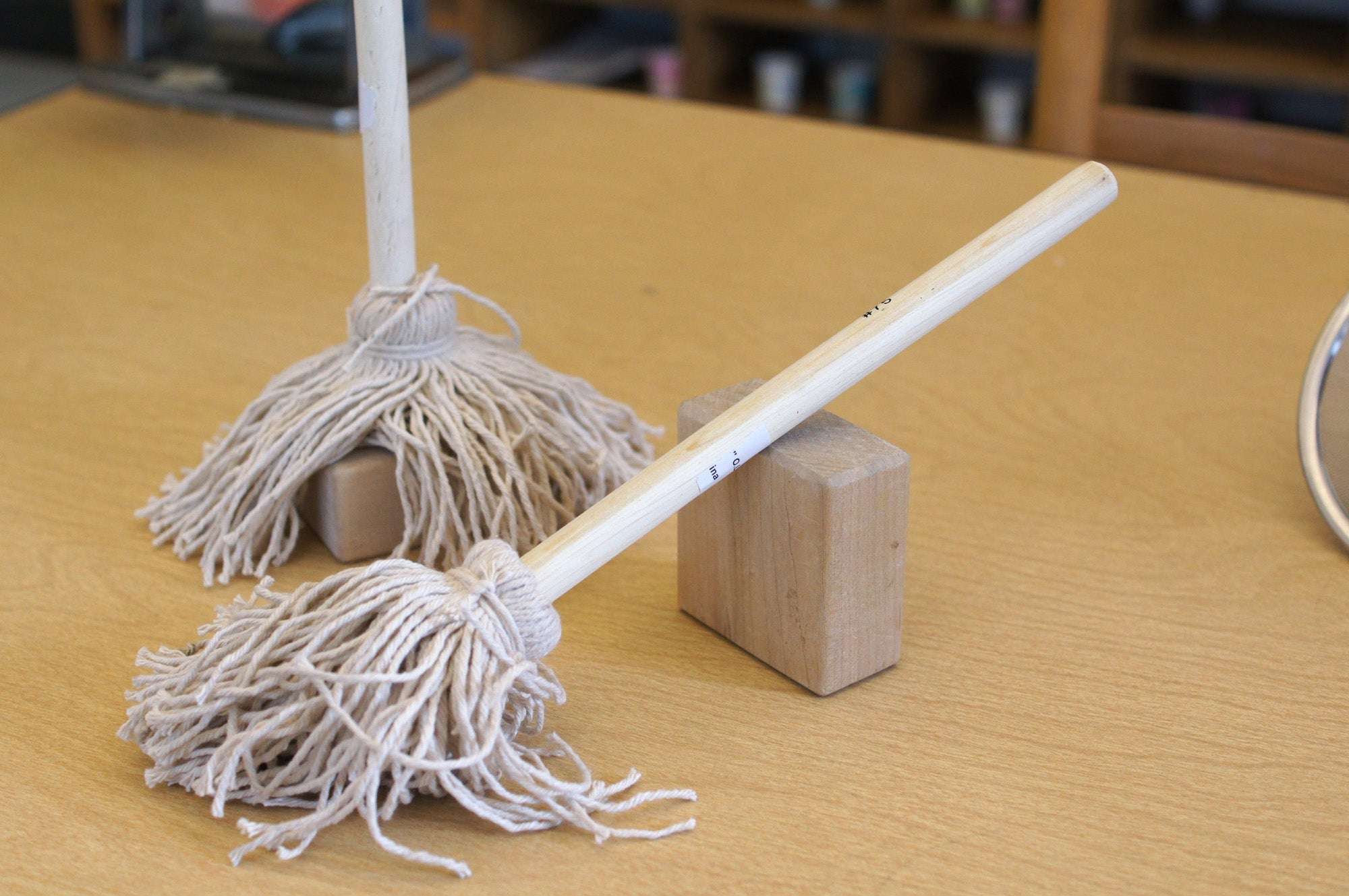 Two mini mops with light wooden handles and thick, natural cotton string heads resting on wooden blocks on a table.