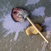 Mini mop with a wooden handle and purple-stained cotton head resting on concrete with pink chalk paint splatters.