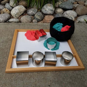 Six stainless steel square and cylindrical molds on a wooden tray with red and blue kinetic sand.