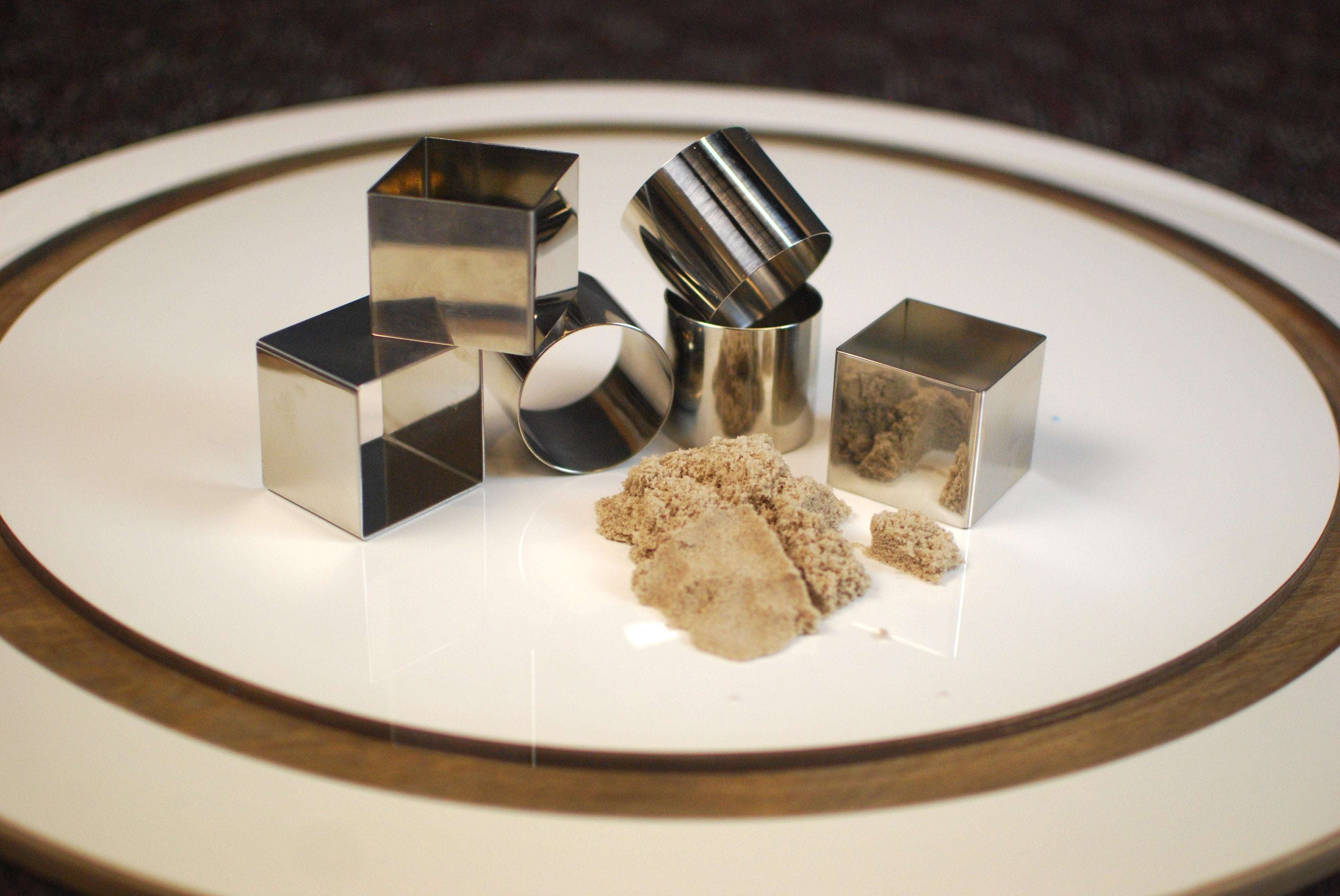 Six polished stainless steel square and cylindrical molds arranged with a pile of beige sensory sand on a white plate.